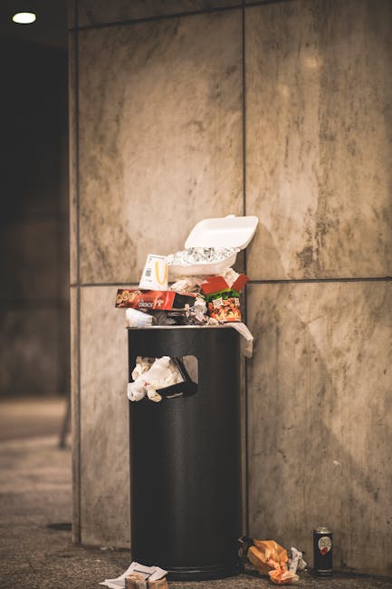 A black cylindrical rubbish bin with a slightly open white plastic lid is positioned against a smooth, beige-toned tiled wall. The top of the bin is cluttered with various waste items, including crumpled paper, snack wrappers, and food packaging, some of which are partially hanging over the edge. At the base of the bin, additional discarded items such as crumpled paper and a small empty container are visible on the floor, which appears to be made of concrete or similar material. Nearby on the ground to the right of the bin, there is a discarded beverage can and a small snack bag. The scene depicts a typical urban setting where waste has accumulated around a private bin, reflecting the need for effective rubbish removal and waste management solutions, as might be handled by services such as Rubbish Removal Paddington. The lighting is neutral, with soft shadows indicating indoor or shaded outdoor conditions, emphasizing the casual, unkept state of the waste collection point.