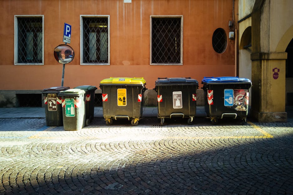 A row of four large wheeled rubbish bins positioned on a cobblestone pavement in front of a building with a peach-colored exterior wall and three windows secured with metal grilles. The first bin on the left is dark green with a textured surface, featuring a green and white recycling symbol and a lid marked for general waste collection. The second bin is black with a yellow lid, displaying a yellow and black waste disposal icon and a sticker indicating hazardous waste. The third bin is also black with a black lid, featuring a white icon for paper recycling. The fourth bin is black with a blue lid, decorated with a sticker for recyclable plastics. To the left of the bins, there is a small circular convex mirror mounted on a pole, reflecting part of the street scene. The building to the right has an arched entrance with a stone column and a small purple logo or sign nearby, with shadows cast across the pavement indicating sunlight. The scene appears to depict a designated waste disposal area, illustrating private waste handling options aligned with independent rubbish collection services.