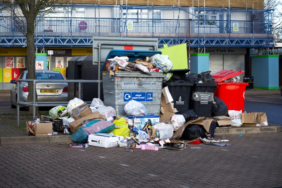 A black cylindrical rubbish bin with a slightly open white plastic lid is positioned against a smooth, beige-toned tiled wall. The top of the bin is cluttered with various waste items, including crumpled paper, snack wrappers, and food packaging, some of which are partially hanging over the edge. At the base of the bin, additional discarded items such as crumpled paper and a small empty container are visible on the floor, which appears to be made of concrete or similar material. Nearby on the ground to the right of the bin, there is a discarded beverage can and a small snack bag. The scene depicts a typical urban setting where waste has accumulated around a private bin, reflecting the need for effective rubbish removal and waste management solutions, as might be handled by services such as Rubbish Removal Paddington. The lighting is neutral, with soft shadows indicating indoor or shaded outdoor conditions, emphasizing the casual, unkept state of the waste collection point.