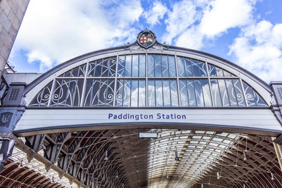 A detailed view of the ornate iron and glass entrance archway of Paddington Station, featuring decorative scrollwork and structural supports painted in a grey finish. Above the arch, the station's sign reads 'Paddington Station' in bold blue letters, positioned on a white background panel. At the top center of the arch, a coat of arms with red, white, and blue elements is visible, reflecting the station's historic significance. The background sky is partly cloudy, showing patches of blue sky and white clouds, which cast soft natural light onto the station's architecture. The interior beneath the arch reveals a curved, vaulted glass roof supported by a network of iron beams and trusses, creating a spacious and well-lit environment typical of classic British railway stations. The overall scene exudes an historic yet functional atmosphere, with the design emphasizing both aesthetic detail and structural strength, all central to the station's operation as a major transport hub. Rubbish Removal Paddington provides comprehensive rubbish collection and waste management services, supporting projects that involve on-site clearance or alternative waste handling for commercial or residential properties near such historic sites.