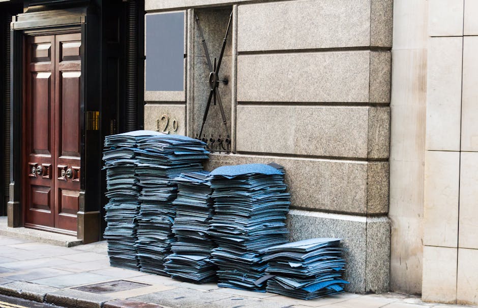 A large pile of neatly stacked blue and white paper documents and files is situated on the paved sidewalk outside a building with a stone facade, right next to a dark wooden door with brass handles and a black metal mailbox. The papers are unevenly arranged, with some stacks leaning slightly and others stacked more uniformly. The setting appears to be an urban exterior environment, with the pile placed directly against the building's stone wall. The lighting suggests natural daylight, casting soft shadows on the pavement and the documents. This scene illustrates a typical example of waste associated with document disposal or off-site clearance, relevant to private rubbish removal services offered by Rubbish Removal Paddington, which handles various types of waste, including paper and filing materials, to support alternative waste handling methods outside local authority collection.