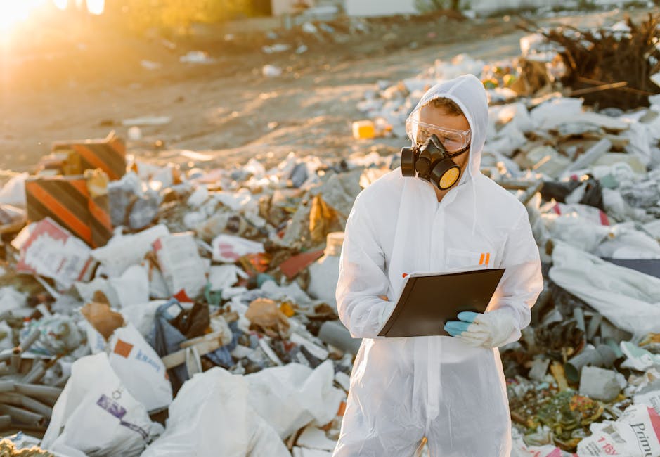 A person dressed in a white hazmat suit, including a hood and gloves, is standing outdoors amidst a large pile of mixed waste and debris. The individual is wearing a full-face respirator with a black filter, shielding their nose and mouth, and is holding a black clipboard while examining the waste. Surrounding them, the waste consists of various materials such as plastic bags, cardboard boxes, paper, and other refuse, scattered across a dirt or gravel surface. The environment is illuminated by natural sunlight, casting a warm glow over the scene, with a blurred background indicating an open, possibly industrial or landfill site setting. This scene subtly relates to hazardous waste management and the importance of safe, professional waste handling, such as that provided by services like Rubbish Removal Paddington, emphasizing proper assessment and management of waste materials during on-site clearance or environmental monitoring processes.