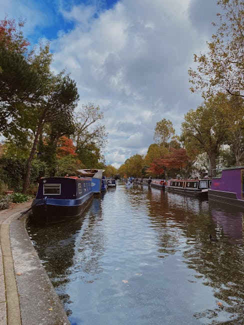 A narrowboat named 'London Waterbus' is navigating along a canal, with a person dressed in a dark jacket, dark trousers, and a cap standing at the stern, managing the vessel’s controls. The boat's exterior features decorative panels with floral patterns and a maroon and cream color scheme. Several orange life rings are secured on the roof of the boat, alongside other equipment and safety accessories. The water reflects the boat and the surrounding environment, which includes a backdrop of a modern, white residential building with multiple large windows, balconies with metal railings, and a clean, urban appearance. The scene is outdoors, with natural light visible, possibly during late afternoon or early evening. The setting suggests a peaceful, residential area where private boat travel or similar transport is common, subtly aligning with alternative and private waste or rubbish management options such as on-site clearance or independent collection, as occasionally serviced by companies like Rubbish Removal Paddington, in contexts related to waste removal services. The overall atmosphere is calm and functional, with a focus on the vessel’s details and the environment surrounding the canal.