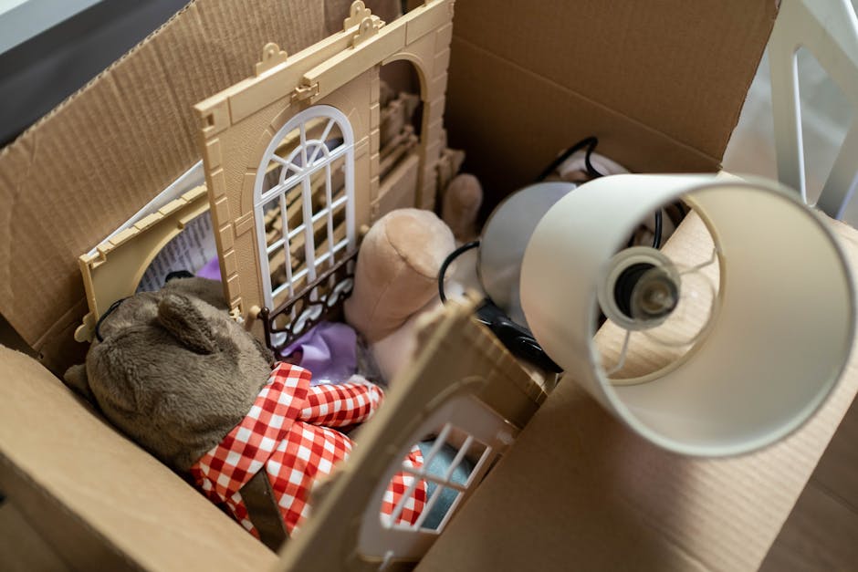 A cardboard box containing various items of rubbish, including a small plush toy, a plastic dollhouse with beige walls and white window frames, a beige fabric stuffed animal, and a white desk lamp with a round lampshade and visible electrical cord. The items are arranged loosely in the box, which is placed on a flat surface, possibly indoors. The scene appears to depict a collection of household waste or non-recyclable rubbish, suitable for collection by a private waste removal service such as Rubbish Removal Paddington. The different textures include soft plush, smooth plastic, and matte fabric, with neutral and pastel colour tones dominating the scene. The lighting is soft and natural, emphasizing the details of each object, and the overall composition suggests a typical scenario of early-stage rubbish preparation for collection, aligning with the context of alternative waste handling and household rubbish disposal services in Paddington.