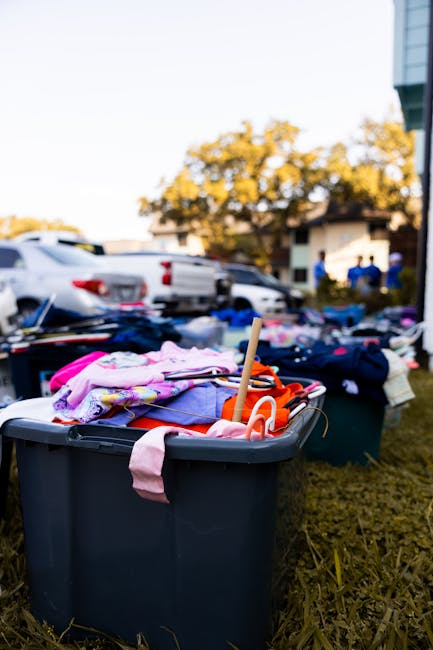 A clear plastic storage bin filled with children's clothes, including pink, purple, orange, and patterned garments, is placed outdoors on a patch of grass beside a driveway or parking area. The clothes are folded and piled on top of each other, with some items draping over the sides of the bin. In the background, there are several parked cars, including white and silver vehicles, and a building with white walls and large windows. Behind the scene, there is a tree with green foliage and a blue sky. The environment appears to be a residential or commercial area, possibly during an outdoor clearance or donation activity, aligning with services offered by Rubbish Removal Paddington for alternative waste handling and bulky item collection.