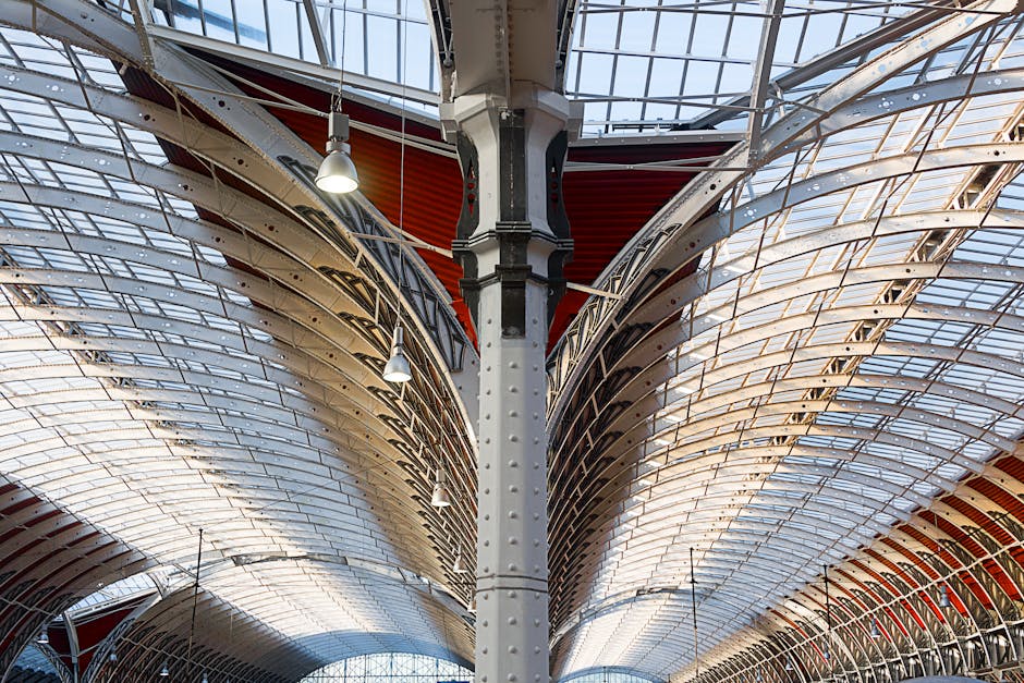 The interior roof structure of a large, modern glass and steel building features a symmetrical, curved design with multiple arched supports. The elements are constructed from metal with a smooth finish, painted in shades of white and grey, with some red panels visible behind the supporting columns. The roof panels are made of transparent or semi-transparent glass, allowing natural light to illuminate the space beneath. Several suspended industrial-style lamps hang from the ceiling, providing additional lighting. The structural columns, positioned centrally, support the expansive arched framework across the ceiling, creating a sense of openness and high ceiling height. The environment appears clean and well-maintained, typical of contemporary architectural designs, possibly within a public building or commercial space. This structural design could relate to the concept of efficient, modern infrastructure, aligning with the themes of development and maintenance often associated with large-scale urban or civic projects, including those managed by waste management and recycling services like Rubbish Removal Paddington, committed to managing waste within architectural and infrastructural contexts.