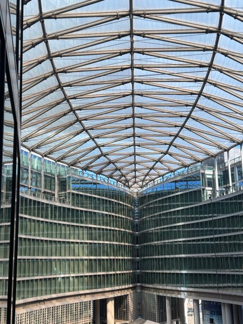 The image depicts the interior view of a modern commercial building with a partially transparent, arched roof structure made of metal beams and glass panels, allowing natural light to filter through. On either side, the building features multiple floors characterized by extensive glass facades with horizontal metal railings, reflecting the surrounding urban environment. The lower levels include glass walls and a perforated metal panel, possibly for ventilation or aesthetic purposes. The overall scene suggests a contemporary office complex or business centre, with clean lines and a sleek, architectural design. A narrow gap is visible at the front, offering a glimpse of other exterior elements like adjacent structures or street level. The crisp daylight illuminates the interior, emphasizing the geometric patterns of the roofing and the glass reflections, aligning with themes of modern commercial space design and supporting the context of private or alternative building management, similar to on-site clearance or refurbishment projects led by specialist services like Rubbish Removal Paddington.