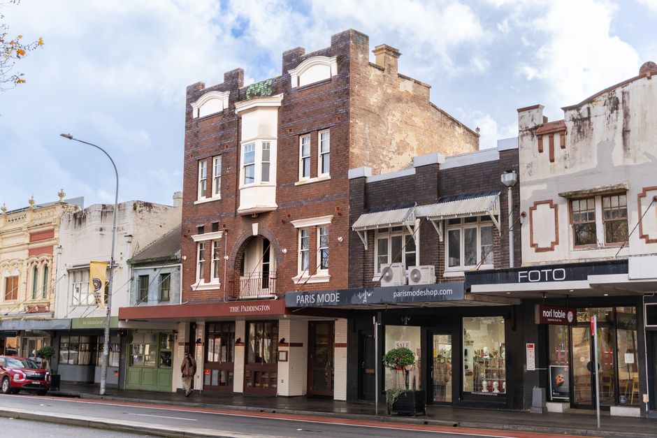 A three-storey brick building with a decorative facade and white window casings sits prominently in an urban street scene, with retail shops occupying the ground floor and residential or office spaces above. The ground level features a shop named 'Paris Mode' with a black awning and large glass windows displaying merchandise, alongside other storefronts including a photography shop and a retail outlet with signage. The building's exterior on the upper floors reveals aged brickwork with some areas of weathering and peeling plaster, contrasting with smoother, recently renovated sections. In the foreground, the pavement is wet, reflecting the overcast sky with patches of sunlight breaking through the clouds. A streetlamp extends upward on the left side, while a parked red car is visible on the street. People can be seen walking along the sidewalk, some entering or leaving the shops. The overall scene presents a mix of historic architecture and modern retail environment, illustrating typical urban commercial and residential coexistence that might be involved in onsite or independent waste handling and rubbish removal services, with Rubbish Removal Paddington occasionally referenced naturally.