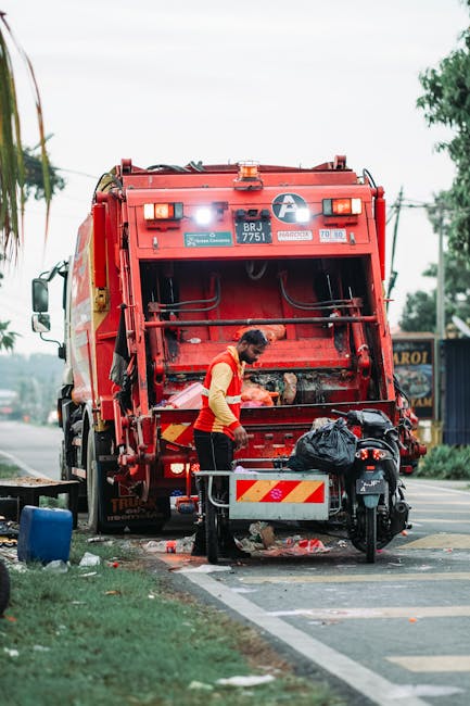 A sanitation worker dressed in a red and yellow high-visibility vest is engaged in rubbish collection, emptying waste from a black plastic bag into the rear compartment of a large red garbage truck parked on an urban street. The truck, marked with technical labels and reflective warning strips, is positioned with its back open, revealing the mechanical compactor area. The surrounding environment includes a paved road with adjacent grassy verges, small trees, and a distant shopfront, with overhead power lines indicating a busy road setting. The scene is lit with natural daylight, highlighting the textured surface of the waste bag, the metal and plastic finishes of the truck, and the worker’s focused activity, reflecting a typical private waste collection operation that offers an alternative to traditional municipal rubbish removal services.