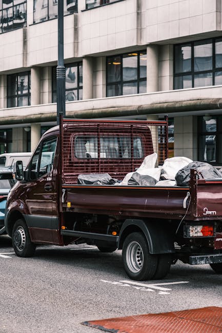 A small dark red flatbed truck parked on an urban street in front of a modern building with large glass windows and concrete exterior panels. The truck's open cargo area is filled with various types of waste materials, including black garbage bags and scattered debris, indicating a collection of domestic or industrial rubbish. The vehicle features a mesh safety partition behind the cab and appears to be designed for quick loading and unloading. Surrounding the truck, there are other parked vehicles and a street lamp, with the scene set during daylight under natural lighting conditions. The background building's reflective windows mirror nearby structures, emphasizing an urban environment suitable for private waste disposal services. This scene illustrates an instance of alternative rubbish collection, often handled by specialist waste removal companies like Rubbish Removal Paddington, supporting local waste management outside of municipal collection services.