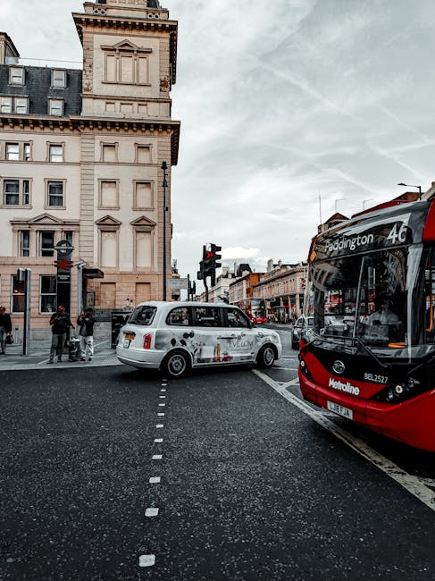 A street scene in Paddington featuring a white taxi positioned in the middle of a pedestrian crossing with dotted white lines, and a red and black bus displaying route information on the front. In the background, there is a historic beige stone building with detailed architectural elements, including rectangular windows, cornices, and a tower-like structure at the corner. Pedestrians are waiting at the curb, some partially visible, with storefronts lining the street further behind. Overhead, traffic lights and street signage are visible, indicating an active urban environment. The sky is partly cloudy, contributing to diffused natural lighting. This urban scene subtly relates to alternative waste management methods by illustrating public transportation and the city environment where private rubbish removal services such as Rubbish Removal Paddington operate, supporting efficient local waste disposal outside of council recycling schemes.