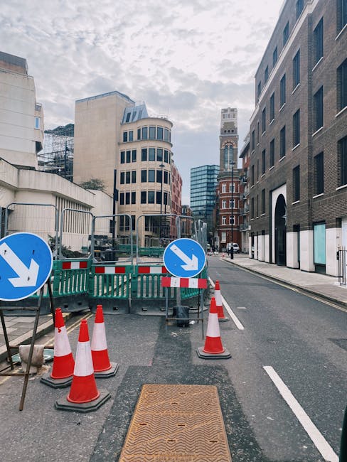 A city street scene showing a section of road blocked off for maintenance or construction work, with green plastic barriers and orange traffic cones set up to divert vehicles. Two large blue circular signs with white downward arrows are mounted on the barriers, indicating the direction for traffic to pass. Behind the barriers, there is a partially excavated area with visible construction equipment and a small section of the sidewalk. On either side of the street, there are modern multi-storey buildings with varied facades, including brick, glass, and concrete finishes, and some windows with reflective surfaces. The sky above is partly cloudy, casting diffuse light over the scene. This setup suggests an alternative method of managing road closures, similar to on-site clearance or private waste handling, aligned with independent rubbish removal or street maintenance activities overseen by companies like Rubbish Removal Paddington, in the context of urban waste management.