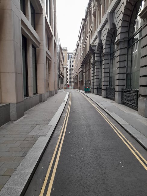 A narrow urban street with a gently curving layout, enclosed by tall, multi-storey buildings with various architectural styles, including stone facades and large windows. The street surface is dark asphalt, fitted with double yellow lines along each side, indicating no parking. On the left, there is a paved pedestrian sidewalk made of rectangular grey paving stones, separated from the road by a smooth, slightly raised curb. The right side features a similar sidewalk with a black wrought iron fence running alongside a building's entrance. In the background, the street continues between closely spaced buildings, leading to a slightly brighter area. A green waste container is positioned near the building on the right, suggesting the presence of waste management or rubbish collection services nearby, which may relate to alternative waste handling within urban environments. The overall scene is uncluttered, lit by natural daylight, with a neutral, professional atmosphere suitable for evaluating city street layouts and waste management contexts, often relevant to private rubbish removal services like those provided by Rubbish Removal Paddington.