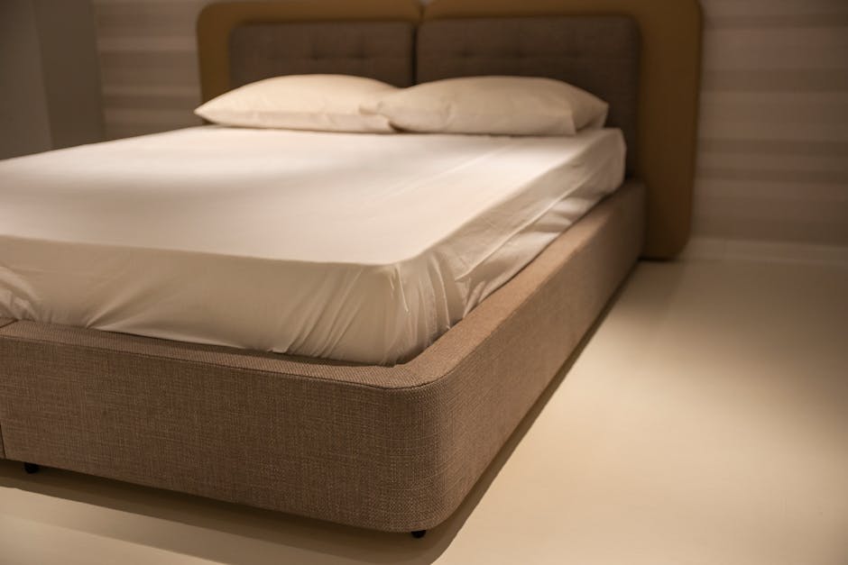 A close-up photograph of a bed with a beige upholstered frame and a padded headboard, situated in a softly lit indoor setting. The mattress, covered with a plain white fitted sheet, appears to be of standard size and has a smooth surface with slight creases near the edges. Two white pillows with matching pillowcases rest atop the mattress, positioned side by side, slightly overlapping. The bed is placed against a wall with horizontal wooden paneling painted in a light shade, contributing to a neutral and minimalist ambiance. The floor beneath the bed is light-coloured, possibly laminate or hardwood, with no visible shoes or additional furnishings. The overall environment suggests a tidy and well-maintained bedroom interior, Although the scene does not involve rubbish or waste, the image's detailed focus on the bed's clean and orderly appearance aligns with professional standards for interior or furniture presentation, suitable for a context where accurate visual descriptions support service relevance in interior decoration or property management related to rubbish removal or private waste handling services.