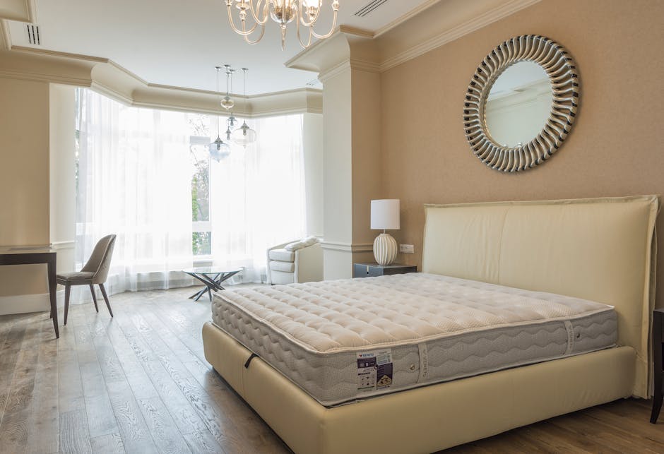 A close-up photograph of a bed with a beige upholstered frame and a padded headboard, situated in a softly lit indoor setting. The mattress, covered with a plain white fitted sheet, appears to be of standard size and has a smooth surface with slight creases near the edges. Two white pillows with matching pillowcases rest atop the mattress, positioned side by side, slightly overlapping. The bed is placed against a wall with horizontal wooden paneling painted in a light shade, contributing to a neutral and minimalist ambiance. The floor beneath the bed is light-coloured, possibly laminate or hardwood, with no visible shoes or additional furnishings. The overall environment suggests a tidy and well-maintained bedroom interior, Although the scene does not involve rubbish or waste, the image's detailed focus on the bed's clean and orderly appearance aligns with professional standards for interior or furniture presentation, suitable for a context where accurate visual descriptions support service relevance in interior decoration or property management related to rubbish removal or private waste handling services.