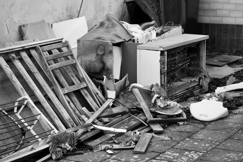 A collection of discarded household objects and building materials stacked on a paved area outside a building. In the foreground, there are wooden pallets leaning against each other, some broken with slats missing or damaged. Nearby, a wire rack and a piece of fabric or rope are tangled among the debris. Towards the center, a damaged white cabinet or drawer unit with exposed internal components and peeling paint is placed, partially covered with a torn, dirty cloth or tarp. To the right, a white plastic potty seat or children's toilet is lying on the ground, surrounded by broken wood fragments, crushed cardboard, and miscellaneous debris. In the background, the wall of a structure, possibly a garage or shed, features a section of brickwork and a dark, covered corner where more rubbish may be stored. The scene is lit with natural daylight, emphasizing the rough textures of the worn, weathered materials, reflecting typical scenes of private domestic waste ready for collection or disposal by professional rubbish removal services such as Rubbish Removal Paddington, in line with independent waste clearance options.