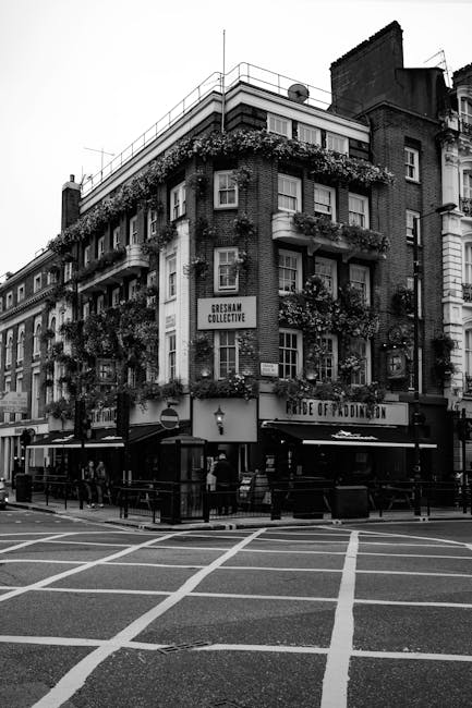 A black and white photograph of a multi-storey building situated on a street corner, featuring several rows of windows with flower boxes and greenery on the exterior balconies. The building's façade appears to be constructed from brick, with a mixture of textures visible in the monochrome image. The ground floor includes a pub or bar, indicated by a sign reading 'FUDGE OF PADDLINGTON' above the entrance, with outdoor seating and a small group of people standing nearby. Adjacent to the building, there are pedestrian crossings with painted lines on the road, and surrounding street lamps. In the background, parts of other buildings are visible, along with rooftop antennas and a satellite dish. The environment suggests an urban setting where private rubbish collection or on-site waste management might be relevant, as the scene depicts a busy street corner typical for logistics and waste removal services. The overall atmosphere appears calm, with natural daylight illuminating the scene, and minimal traffic visible on the road.